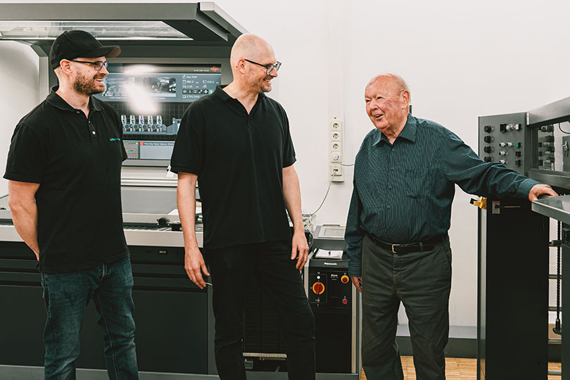 75 Jahre Erfahrung und Werte - Drei Männer stehen zusammen. Der ältere Mann, der Senior der Firma, lächelt den beiden jüngeren Männern zu. Im Hintergrund ist ein großer Leuchttisch zu sehen, auf dem Druckerzeugnisse kontrolliert werden. Der Senior lehnt sich seitlich an eine Druckmaschine.
