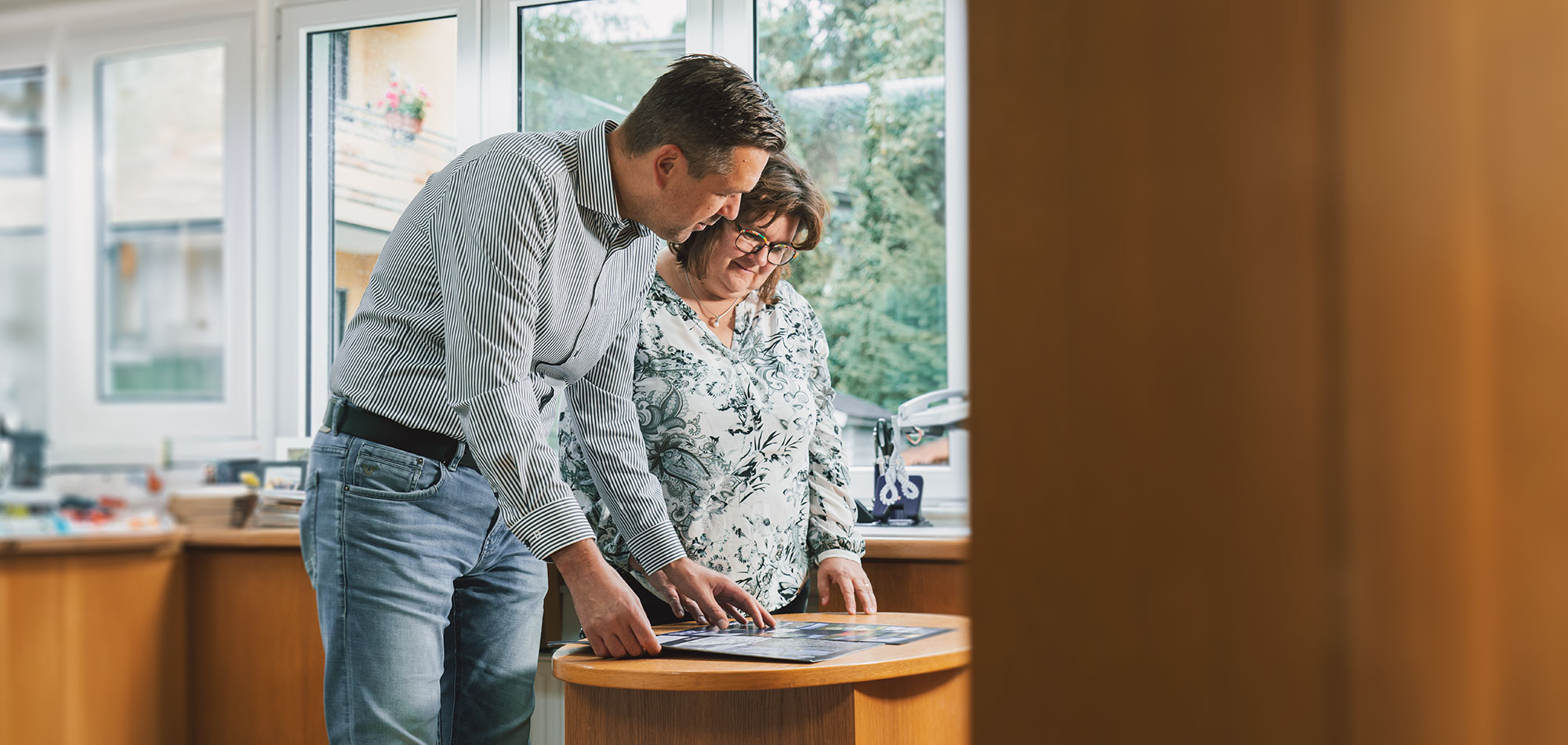 Über uns, Ein Mann und eine Frau stehen und sehen sich ein Druckprodukt an, das auf einem kleinen Tisch liegt. Die Perspektive geht um die Ecke.