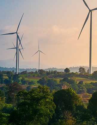 VERANTWORTUNGSVOLLER UMGANG MIT ENERGIE - Eine Windparkanlage mit fünf Windenergieanlagen. Im unteren Bereich des Bildes ist eine grüne Landschaft mit Bäumen und Feldern zu sehen, während im oberen Bereich ein Abend- oder Morgenrot leuchtet.